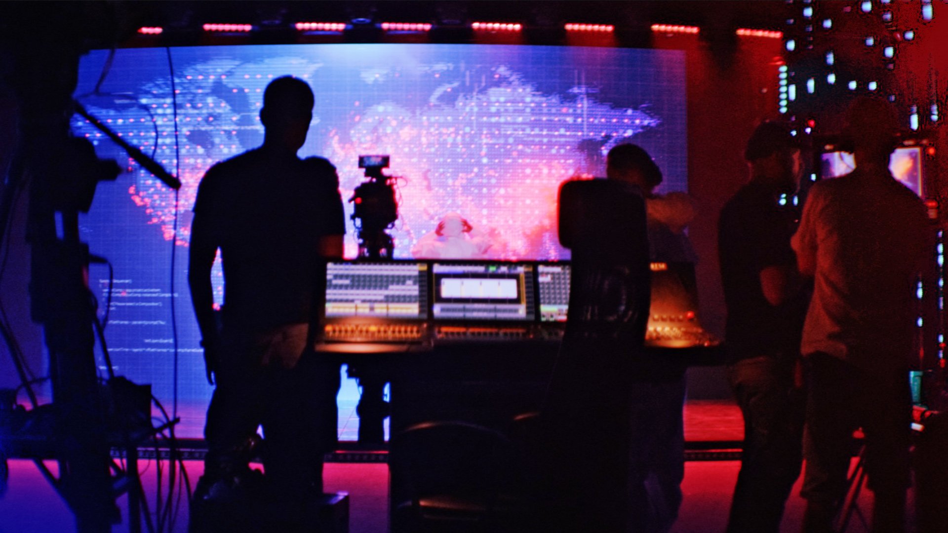 Behind-the-scenes view of a modern news studio featuring backlit audio mixing boards, silhouette of a broadcast crew, and a digital global map display.