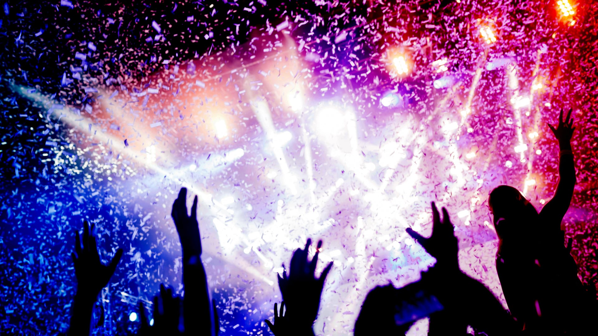 Silhouettes of a cheering concert crowd with raised hands illuminated by vibrant stage lights and falling confetti during a live event.