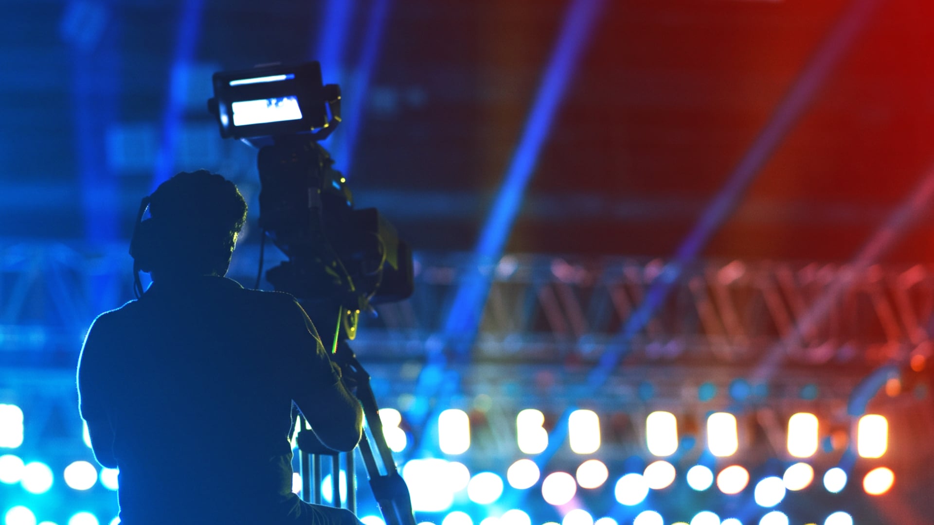 Silhouette of a camera operator filming a live sports broadcast under dramatic blue and red stage lighting.